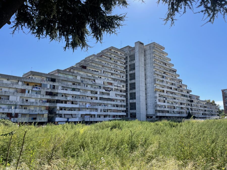 Vela Scampia terraced apartment block with concrete balconies and overgrown foreground vegetation.