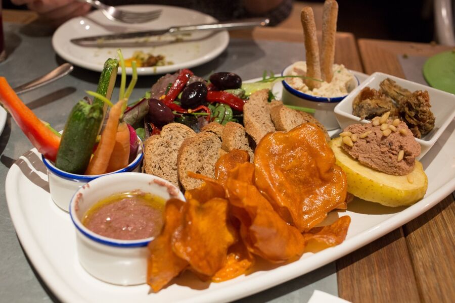 Appetizer platter with sweet potato chips, rustic bread, veggies, dip, marinated olives, and spreads.