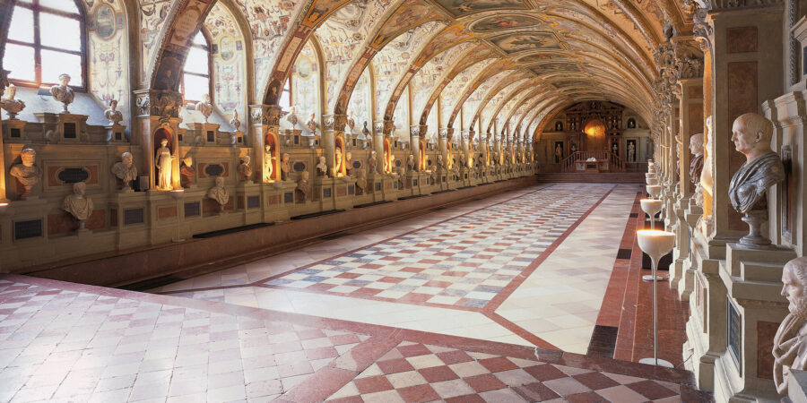 Arched gallery with barrel vault fresco ceiling and marble sculptures in symmetrical museum corridor.