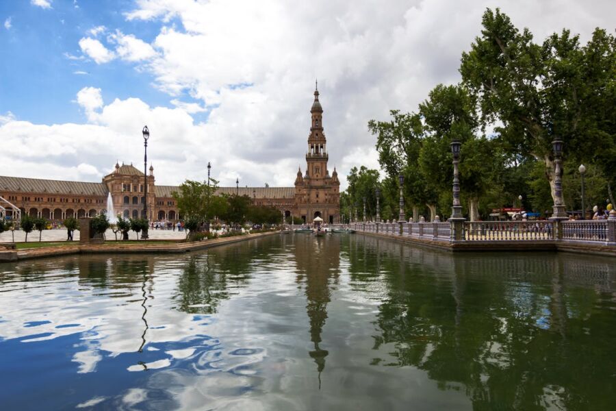 Sunny Spanish plaza with tower, water reflections, boats, and lively crowd amid historic architecture.