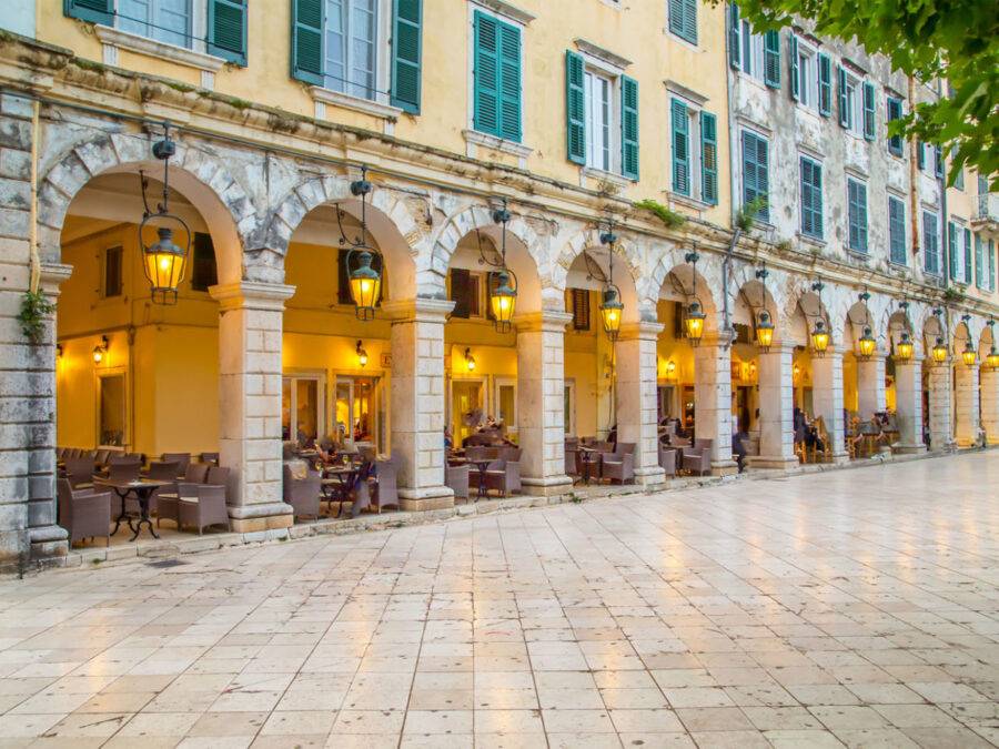 Historic stone arcade with arches, green shutters, cozy seating, and evening lantern glow.