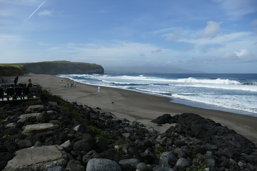 Rocky foreground leading to a sandy beach of Areal de Santa Bárbara in Portugal with waves crashing under a partly cloudy sky. Distant cliffs and people walking create a serene atmosphere.