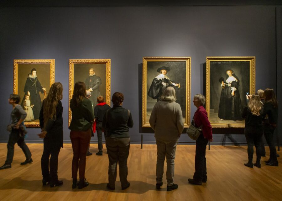 Art gallery with historical portraits, diverse visitors, ornate frames, and deep blue walls.