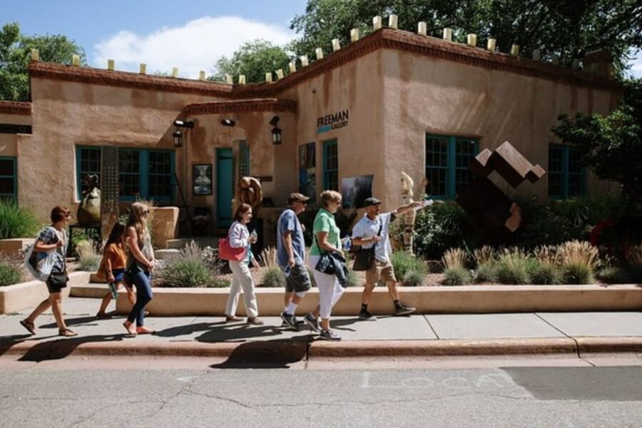 Artistic district scene with people in summer attire outside adobe-style building and sculptures.