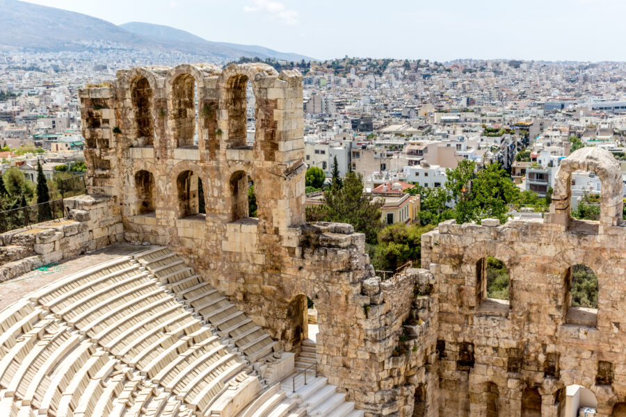 The iconic Athens Theater featuring tiered stone seats and a scenic backdrop of ancient ruins and vibrant foliage