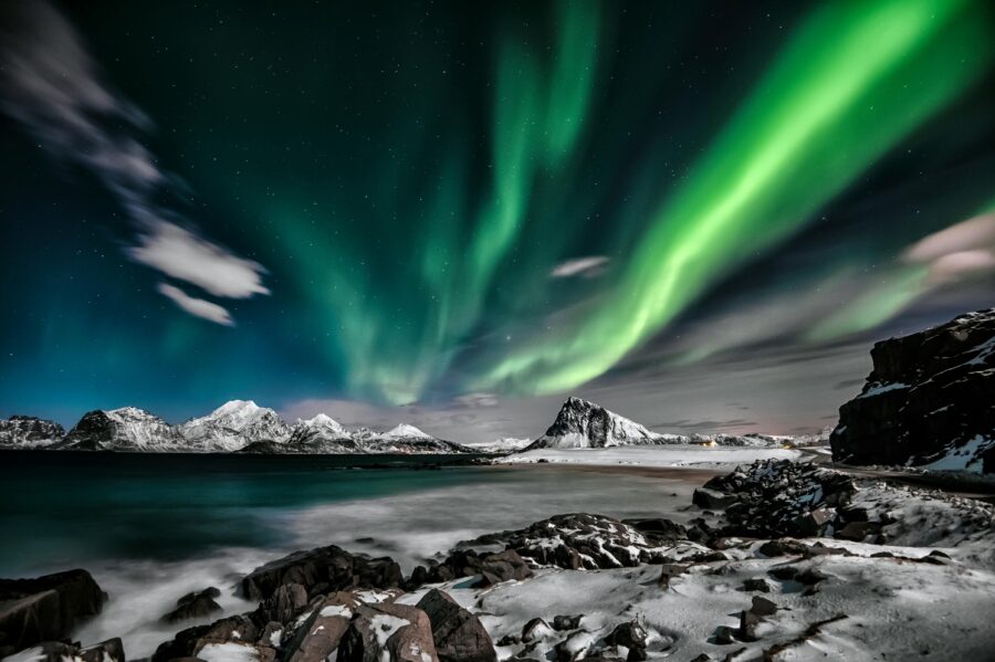 Northern Lights dance above snowy mountains, rocky shores, and a serene sea reflecting vibrant colors.