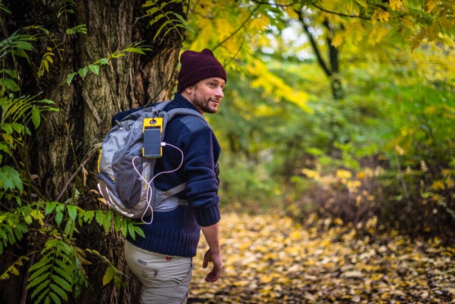 Man in autumn forest with backpack, wearing a beanie, showcasing outdoor adventure and technology.
