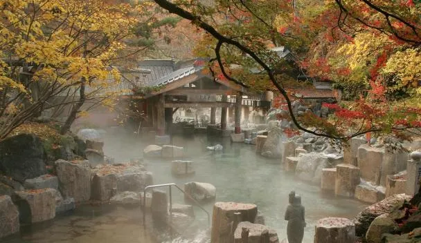 Tranquil Japanese outdoor onsen hot spring in autumn forest with steam, rocks, and wooden bathhouse.