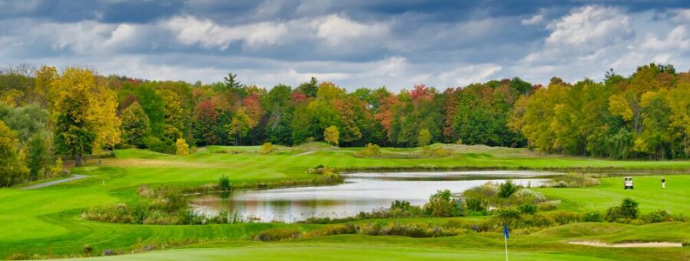 Autumn golf course scene with vibrant foliage, cloudy sky, pond, and golfers playing.