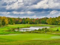 Autumn golf course scene with vibrant foliage, cloudy sky, pond, and golfers playing.