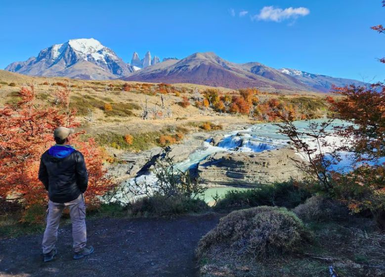 Hiker admires autumn landscape with river, waterfalls, and snow-capped mountains.