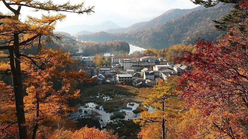 Autumn mountain village by lake, colorful forest trees, scenic Japanese onsen resort landscape.