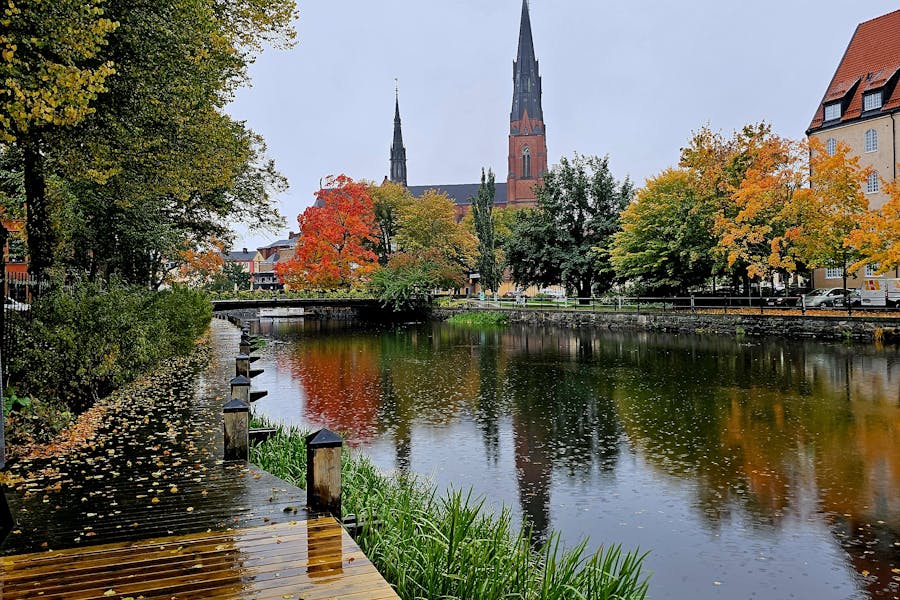 Autumn river view of Uppsala Cathedral spires with bridge, wet boardwalk, and colorful reflections.