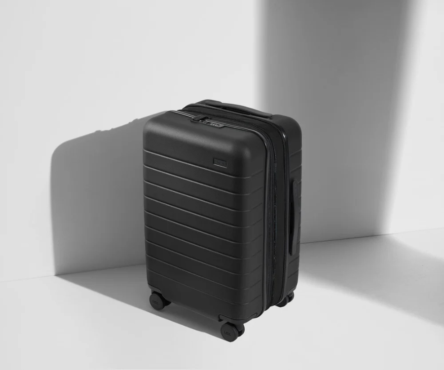Black Away Carry-On Flex suitcase rests on a white surface, casting a soft shadow beneath it.
