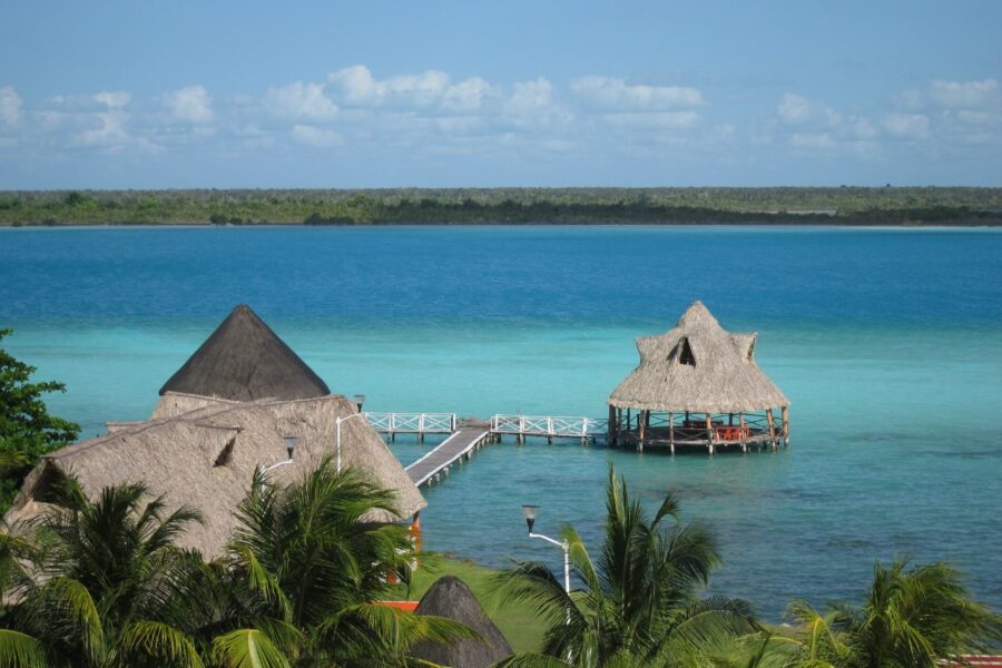 Scenic view of Bacalar Lagoon beach, featuring a pier and thatched huts under a clear blue sky in Quintana Roo, Mexico.