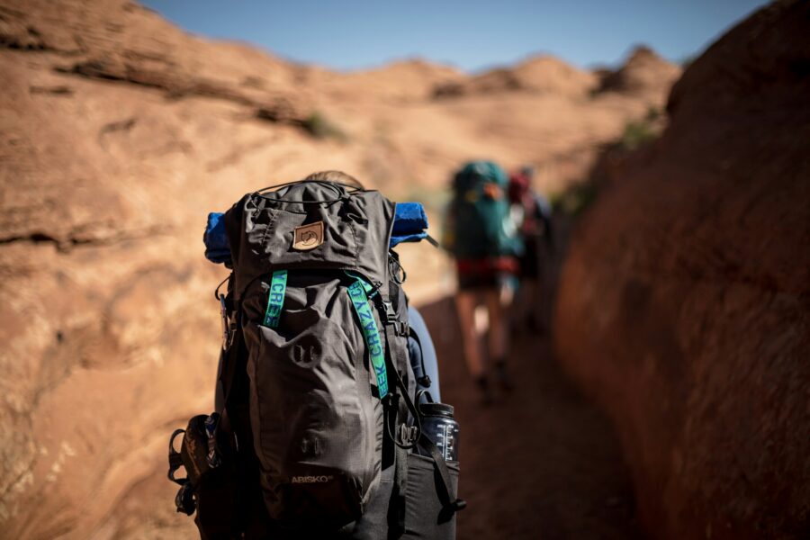 Sunlit slot canyon hikers with foreground backpack and rolled blue sleeping pad.