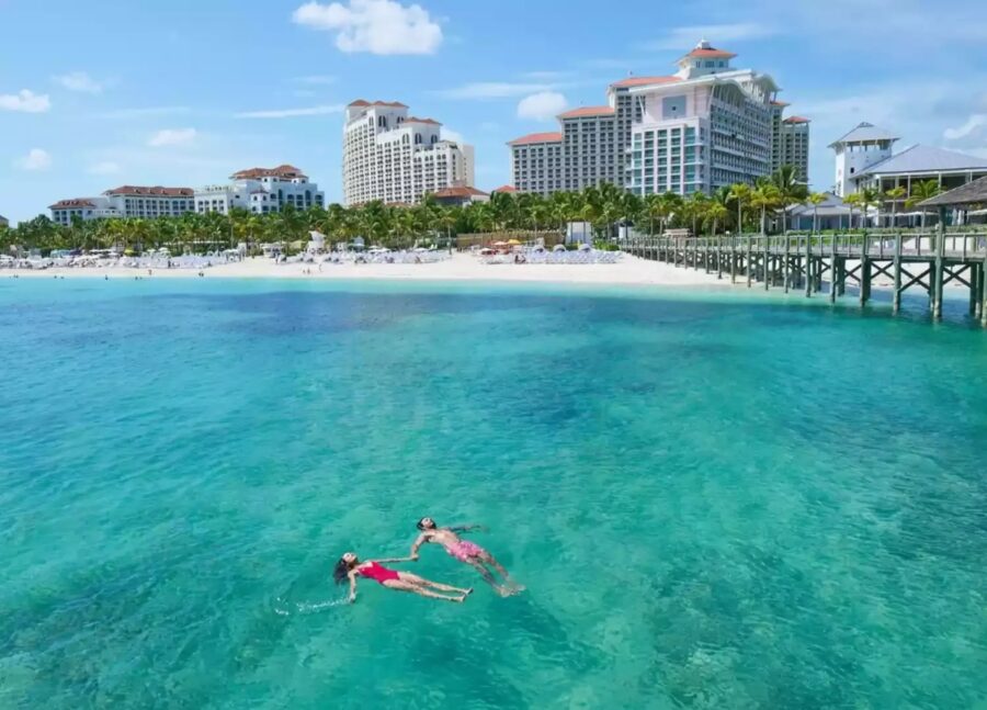 Two people swim in the ocean near a pier at Baha Mar, Nassau, Bahamas, under a clear blue sky.