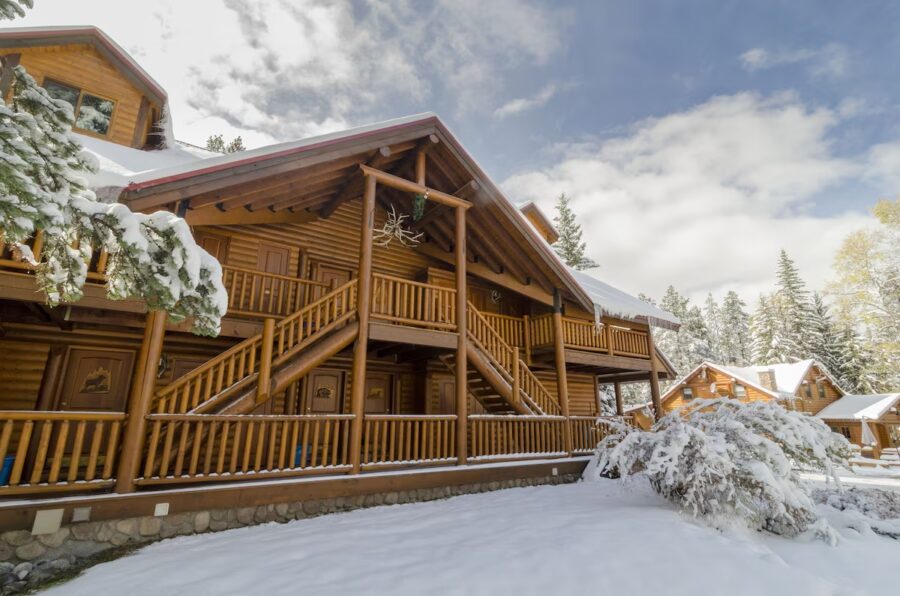 Charming log cabin with a snowy porch and evergreen trees, creating a picturesque winter scene at Baker Creek By Basecamp.
