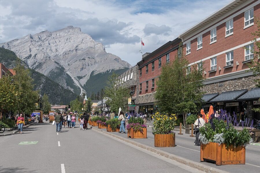People stroll along a street in Banff, Alberta, with a stunning mountain range visible in the background.
