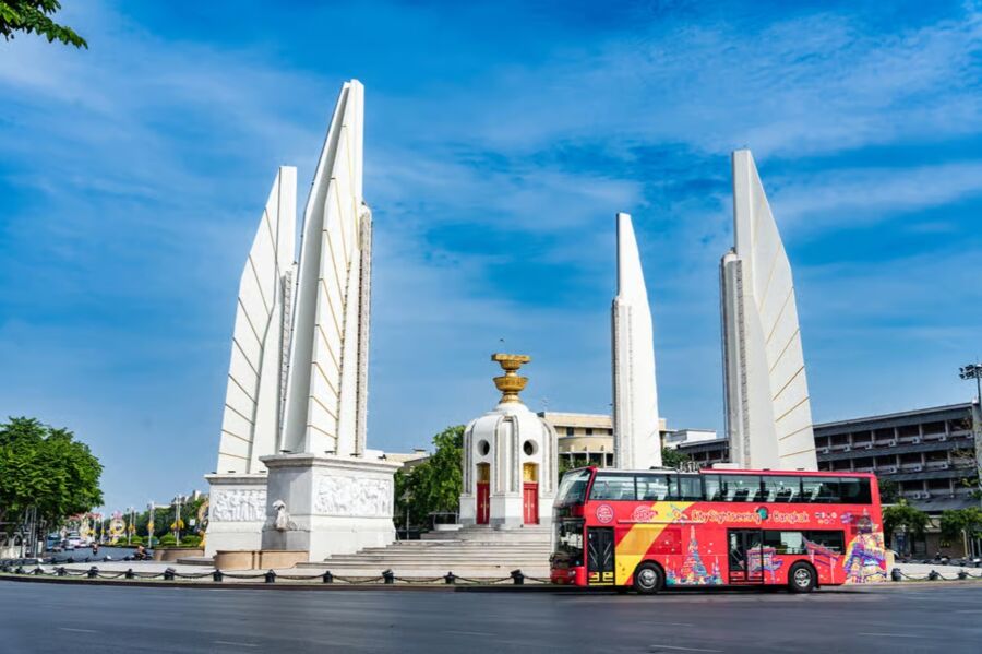 Vibrant red double-decker bus drives past the imposing Democracy Monument in Bangkok under a clear blue sky, capturing a sense of urban energy.