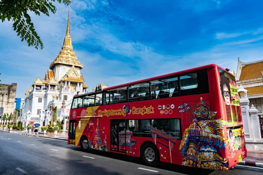 Red double-decker sightseeing bus drives past the ornate, golden-spired Wat Traimit in Bangkok under a clear blue sky, capturing a lively urban scene.