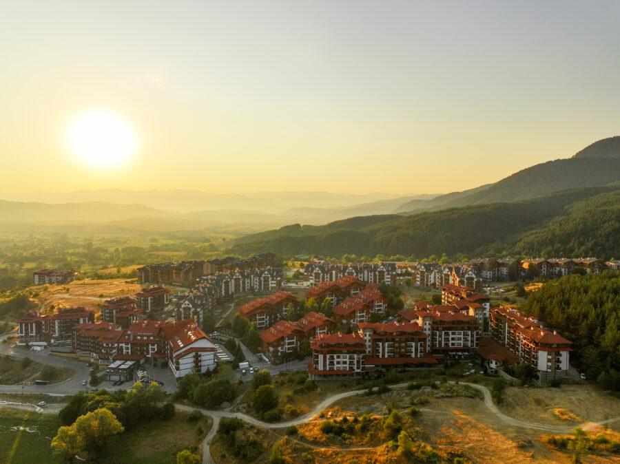 Warm golden hues cover Bansko, Bulgaria, as sun rises over mountain peaks. Aerial view showcases tranquil town, blending traditional architecture with breathtaking natural surroundings.