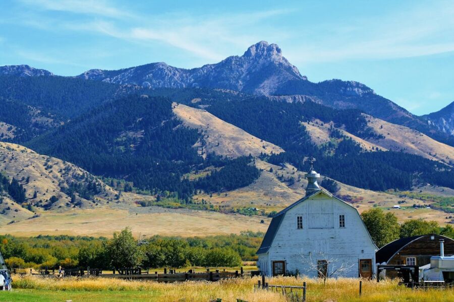 Serene rural scene: white barn, grassy fields, majestic mountains under a clear blue sky.