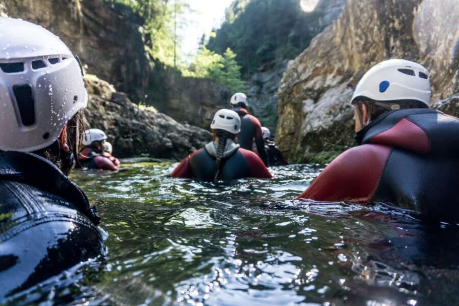 Group of people wearing helmets and wetsuits are canyoning through a narrow, rocky river in Bavaria. Sunlight filters through trees, creating a calm, adventurous mood.
