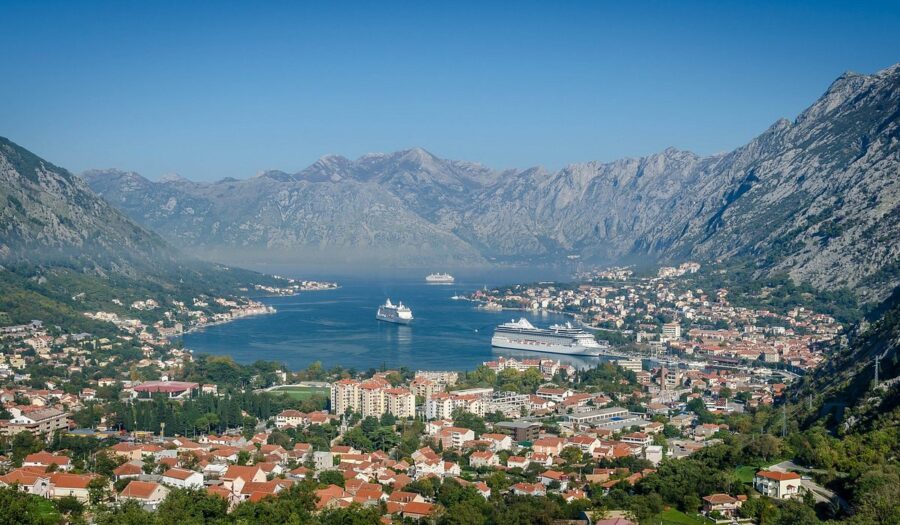 Scenic coastal town with mountains, terracotta rooftops, vibrant harbor, and clear blue skies.