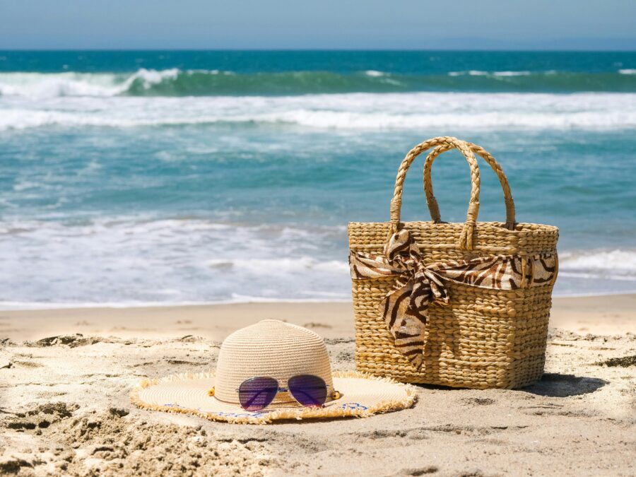 Straw beach bag, hat and sunglasses on sandy shore by blue ocean waves.