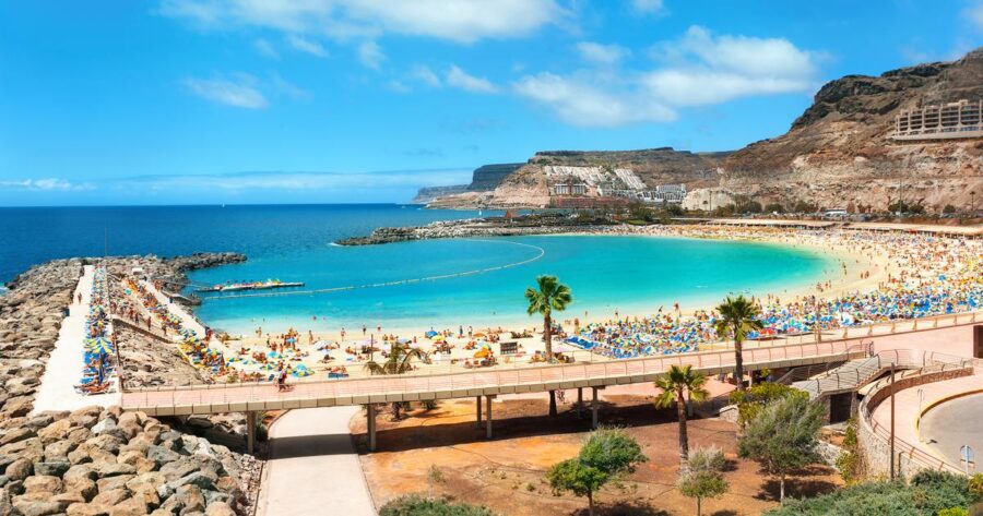Gran Canaria turquoise bay beach with promenade, cliffs, umbrellas and sunbathers.