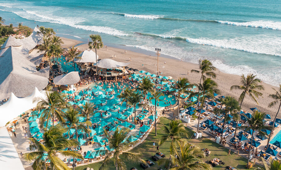 Luxury Bali beach club with oceanfront infinity pools, palm trees, sun loungers, umbrellas, and crowds.