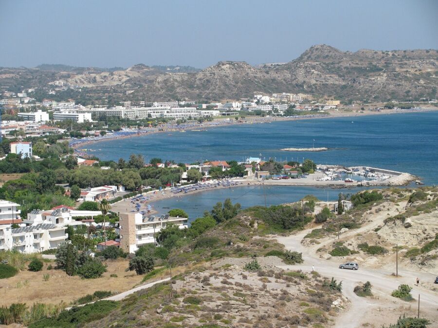 Seaside town with beach, marina, palm trees, and hills under a sunny blue sky.