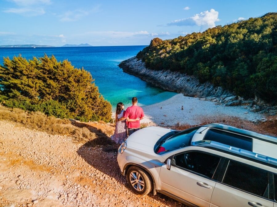 Serene beach scene with couple, turquoise waters, lush greenery, SUV, and distant mountains.