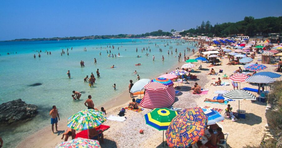 Summer day at Fontane Bianche beach with colorful umbrellas, clear waters, and lively activities.