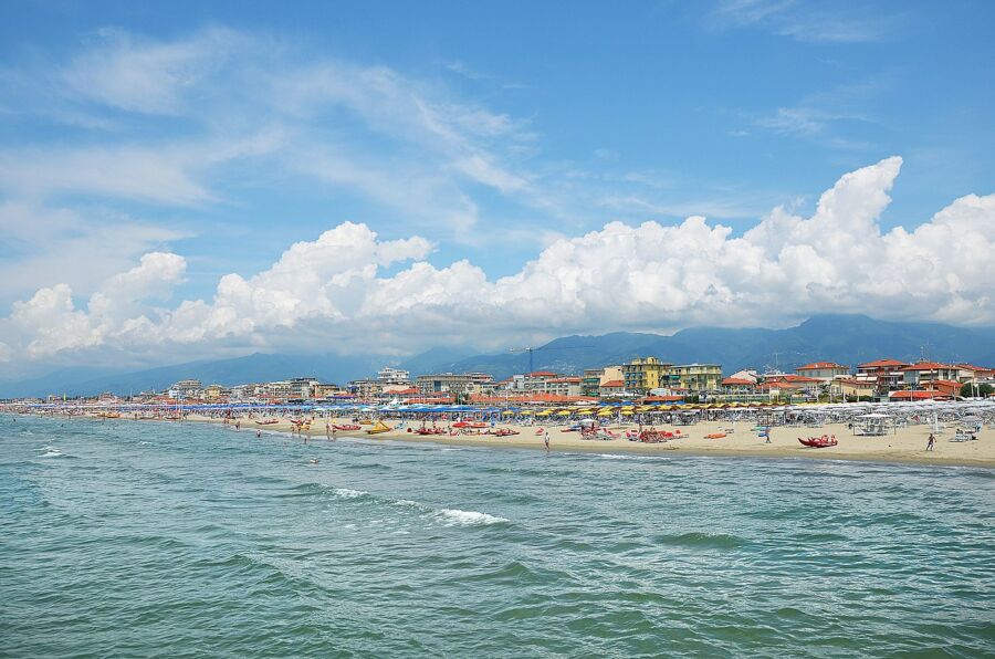 Sunny beach scene with people, umbrellas, ocean, and mountains in Lido di Camaiore, Italy.