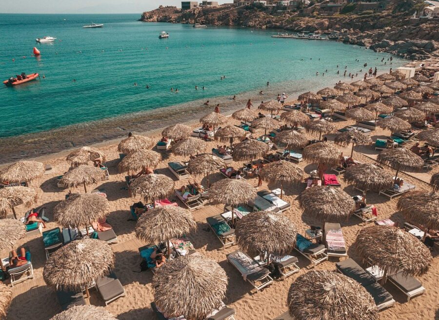 Sunny beach scene with turquoise waters, sunbathers, boats, rocky coast, and palm-thatch umbrellas.