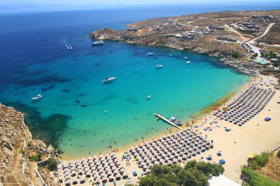 Idyllic Mediterranean beach with turquoise waters, yachts, sunbathers, and picturesque hills.
