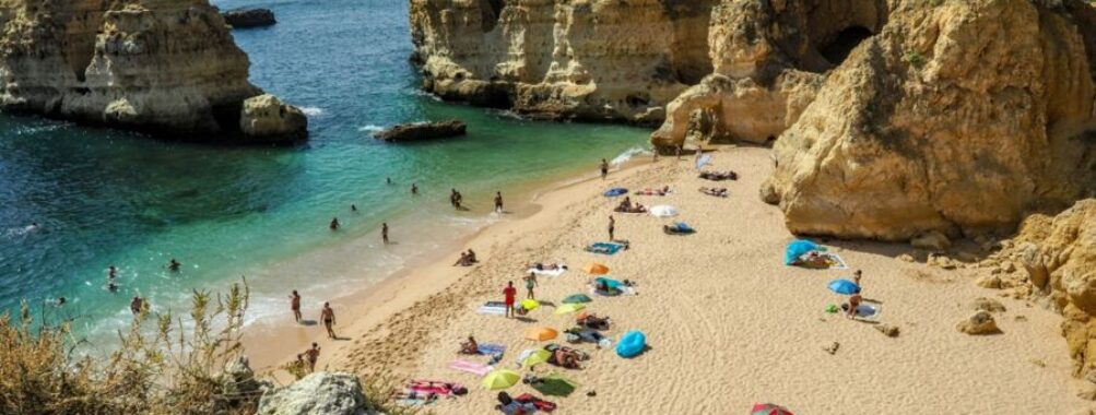 Picturesque secluded Algarve beach with cliffs, turquoise sea, beachgoers, colorful towels, and umbrellas.