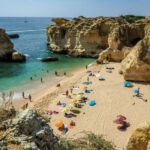 Picturesque secluded Algarve beach with cliffs, turquoise sea, beachgoers, colorful towels, and umbrellas.