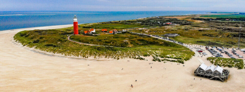 Aerial view of wide sandy beach with red lighthouse, dunes, sea, and coastal parking area.