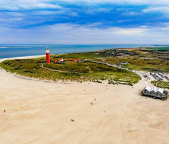 Aerial view of wide sandy beach with red lighthouse, dunes, sea, and coastal parking area.