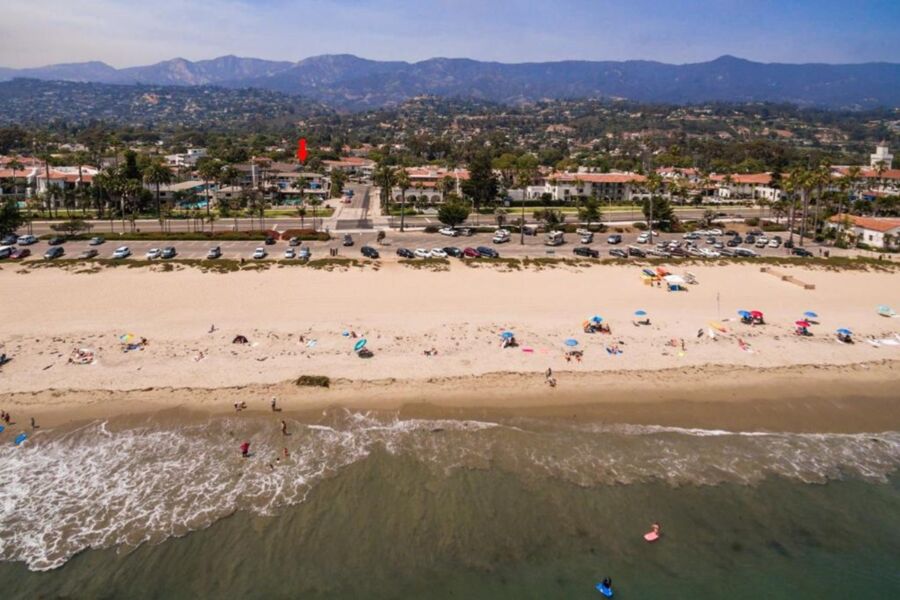 Coastal beach scene with sandy shore, colorful umbrellas, palm trees, mountains, and partially cloudy sky.