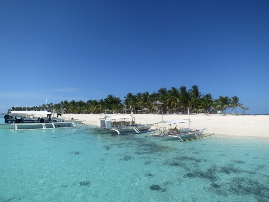 Malapascua beach with outrigger boats, turquoise water, white sand and palm trees.