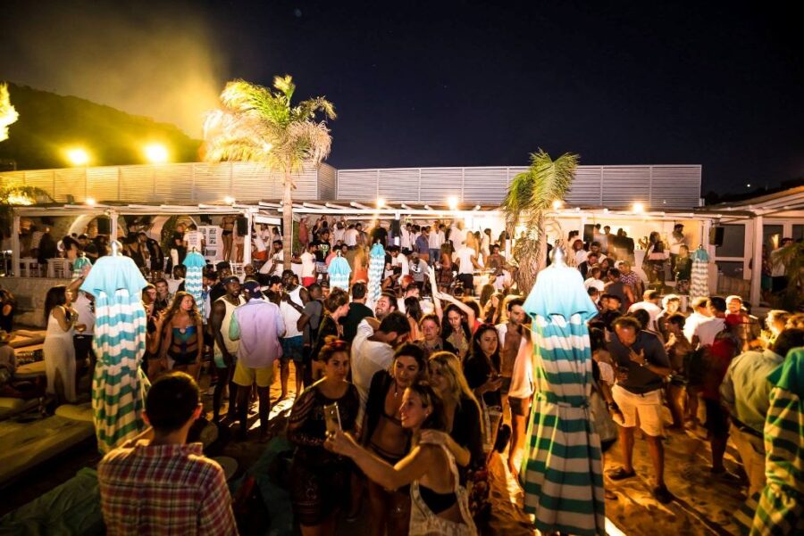 Beach party nightlife under starry sky with music, drinks, lively crowd, and tropical vibes.