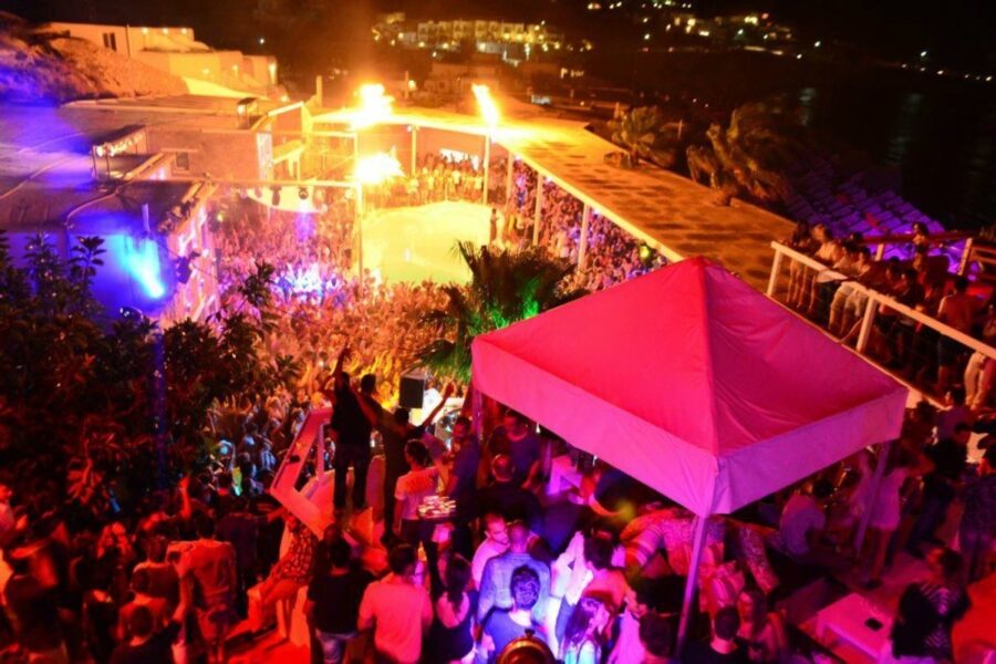 Vibrant beachside party in Mykonos with colorful lights, dancing crowd, and fiery spectacles.