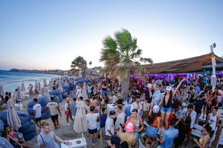 Sunset beach party with diverse crowd, palm trees, music, and vibrant summer atmosphere.