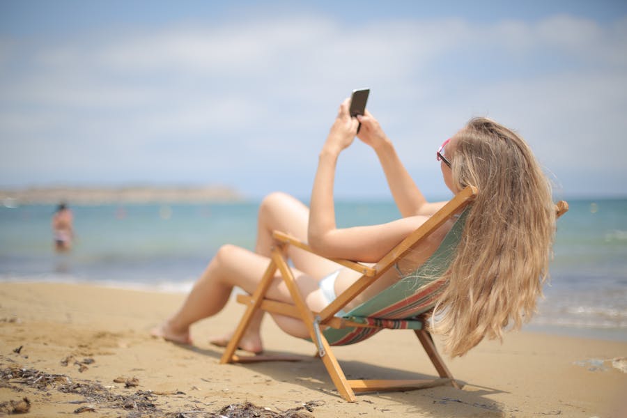 Woman relaxing on beach chair with smartphone, ocean in background.