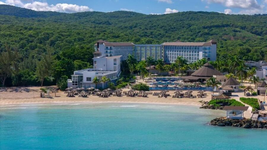 Modern beachfront resort with lush hills, golden sand, turquoise waves, and inviting swimming pool.