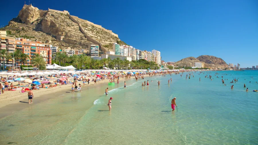 Sunlit beach scene with colorful umbrellas, palm trees, shimmering water, and sandstone mountain backdrop.